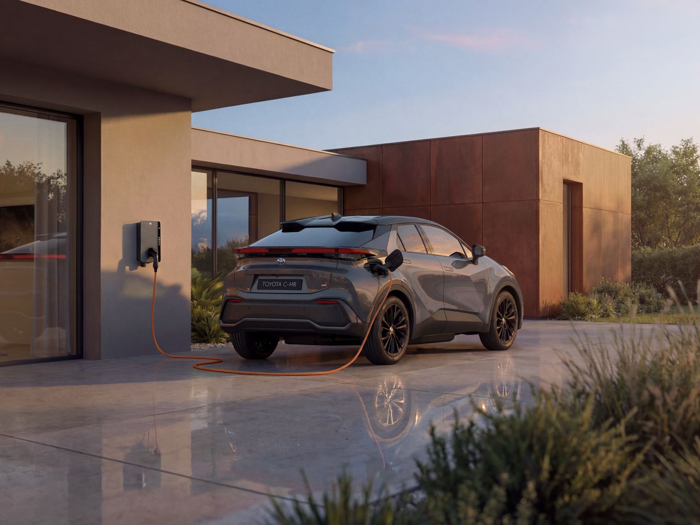 Toyota C-HR Elektroauto in dunkelgrauer Farbe lädt an einer Wandladestation vor modernem Haus mit großen Fenstern und Holzverkleidung